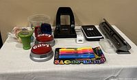 Photo shows a collection of used school supplies arranged on a white cloth, including a black hole punch, scientific calculator, pack of highlighters, red Easy buttons, tape dispensers, and various small office supply containers.