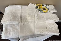 Several folded white lace and embroidered tablecloths stacked on a surface.