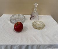 Front view of the three glass items on a white cloth with an apple for scale: decanter with stopper, clear cut glass bowl, and small yellow glass dish.