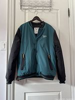Front view of vintage style Reebok jacket hung on door, showing teal body and black sleeves, snap button closure and zippered pockets.