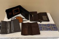 Eight wallets laid flat on a white surface showing various styles including bi-fold, long wallets, and fabric clutch with visible compartments and detailing.