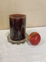 Dark brown wax candle sitting on a clear scalloped glass candle holder next to a red apple for scale.