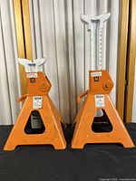 Two orange Allied jack stands of different heights shown against curtain background on black table. Metal construction, adjustable ratchet bar with lever.