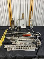 Photo showing a variety of large metal drill bits, sockets, ratchet wrench, and hand saw frame with wooden handle laid out against a black cloth background.