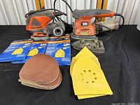Two Black & Decker corded electric sanders with several sanding sheets displayed on table.