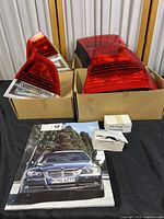Photo showing two red and clear BMW taillight lens assemblies placed in cardboard boxes with three small boxed parts and a BMW 3 Series Sedan brochure/booklet below.
