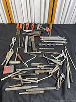 Overview of multiple tools laid out on a black surface including pliers, screwdrivers, drill bits, wrenches, and various metal rods and tools.
