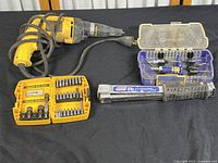 DeWalt DW257 drill with cord, sets of bits in yellow and blue cases, screwdriver extension handle and additional bit holder on black background.