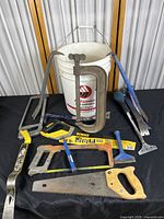 Photo showing a variety of hand tools on a black surface, including pry bars, saws, clamps, scrapers, alongside a white plastic bucket in the background.