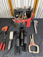 Red and black Husky tool bag with various hand tools on black cloth, three black Maglite flashlights, pipe wrench, locking plier, putty knife, wire crimper, and pipe cutter.