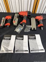 Husky nail tool set displayed on black cloth, showing three air nailers arranged next to each other along with manuals and small accessory parts in sealed plastic bags.