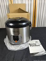 Full view of the Kevin Dundon electric multi-cooker showing stainless steel body, black lid, keypad and included manual beside it with cardboard box in background.