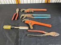 Full view of 6 vintage metal tools including 3 pipe wrenches, 2 pliers, and 1 file on black surface