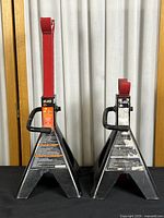 Pair of Team Mechanix 2-ton capacity steel jack stands, black base with red adjustable arms, standing side by side in front of a curtain.