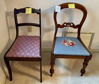 Two antique style needlepoint chairs side by side against a white wall with electrical outlet and vent in background.