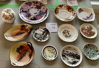 Overview of 12 decorative bone china plates laid out on a table with various floral and scenic designs, some oval and some round in shape.