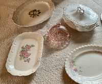 Seven pieces shown: pink depression glass butter dish with lid, white covered casserole dish with silver accents, rectangular floral bone china dish, several floral-patterned platters with scalloped and embossed edges.