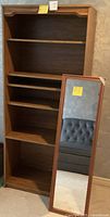 Photo showing wooden bookshelf with four shelves and full-length wall mirror leaning against the bookshelf.