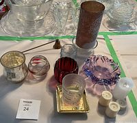 Photo showing a group of various crystal and glass candle holders along with some candles and a metal candle snuffer on a white surface with green tape lines.
