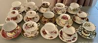Top-down photo showing all 16 pairs of tea cups and saucers, arranged in rows, displaying varied floral and gold designs.