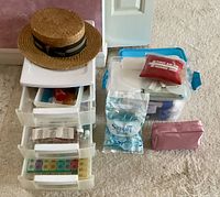 Shows white plastic drawer organizer with first aid supplies and pill organizers on top, along with straw boater hat and blue/clear plastic containers with bandages and pouches.