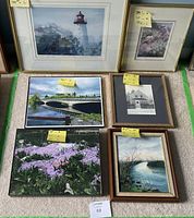 Lot of six framed art pieces arranged on carpet, showing various subjects including landscapes, floral artwork, a lighthouse, and architectural sketches with size notes on yellow sticky tabs.