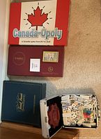 Image of closed Canada-Opoly, Scrabble, and Trivia Pursuit board game boxes stacked on the floor near Train Dominoes case.
