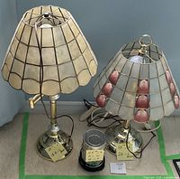 Two vintage brass table lamps with capiz shell lampshades, one cream-colored and one with pink scallop shell accents, shown side by side on floor.