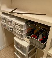 Wide view of white plastic storage drawers and tools in gray bin on shelf with metal carpenter's square level resting above