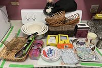 Photo shows multiple kitchen items including ceramic bowls and plates, woven baskets, plastic cutlery packs, paper plates, straws, paper napkins, decorative saucers, and disposable cups arranged on a kitchen stove surface.