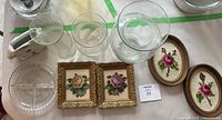 Photo showing a collection of three clear glass vases, two framed needlepoint rose artworks in oval wooden frames, a framed needlepoint floral artwork in a square wooden frame, and a clear divided crystal dish on a white surface.