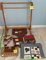 Photo of wooden quilt rack and six assorted quilted textile items arranged on floor including wall hangings and runners in various patchwork designs and colors