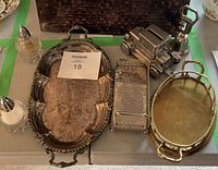 Photo showing two oval silver plated serving trays, one with a scallop rim and the other with smooth gold-tone finish. Also displayed are a rectangular hammered silver tray, a diecast Ford Model T coin bank, a silver bell, and salt and pepper shakers on a white tabletop.