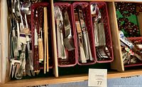 Drawer with several trays containing mixed silverware and serving utensils, plus visible napkin rings to the side
