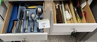 Two kitchen drawers. First drawer contains bread knife, stainless steel forks, spoons, knives, and serving spoons in gray plastic tray. Adjacent drawer holds utensils including ladles, peelers, whisks and other kitchen tools separated by wooden compartments.