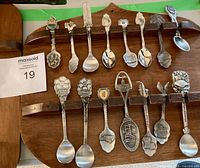 Photo showing 14 souvenir spoons on a wooden rack, each with unique decorative handles depicting flowers, crests, and landmarks.