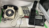 Photo shows four landline telephones on a table: vintage beige rotary phone, cream large-button push-button phone, black Panasonic cordless phone base and handset, plus a separate cordless handset.