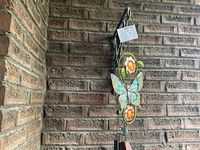 Wind chime with blue butterfly and yellow flower accents hanging on black metal hanger against brick wall.