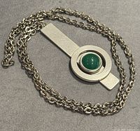 Full view of the pewter necklace with a long chain and circular pendant with green stone on gray background.