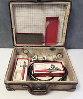 Open case showing two vintage metal airbrush guns with red handles, coiled air hose, small glass jars with lids, and nozzle accessories in compartment trays lined with grid-pattern fabric.