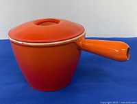 Front and side view of orange enameled cast iron fondue pot with lid and long handle on blue background