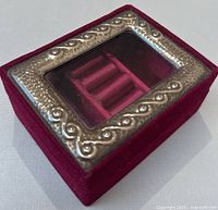 View of the top of the burgundy velvet jewelry box showing silver-tone metal decorative frame with embossed scroll pattern surrounding the clear glass peek window