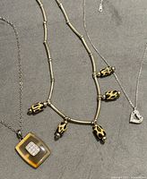 Three silver necklaces displayed on grey fabric; includes a beaded necklace with tan and black patterns, amber-colored square pendant necklace, and heart-shaped rhinestone pendant necklace.