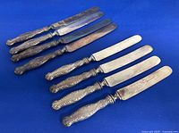 Eight antique knives displayed on blue background, showing ornate silver handles and blades with some wear and patina.