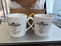 Photo of two white ceramic coffee mugs with black text commemorating 50 years of marriage, featuring heart-shaped handles forming a heart when placed together.