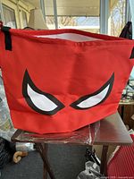 Photo of a red fabric laundry bag with black and white angry eye graphic design on the side, showing black handles and an open top.