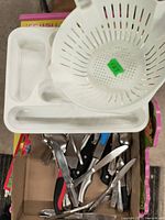 White plastic cutlery tray and white colander placed on top of a box with assorted kitchen tools visible below.