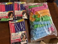 Photo showing several packaged sets of soft foam hair curlers in pink and purple, along with a plastic bag containing loose colorful wave-shaped curlers.