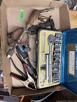 Box containing a vintage hand-crank drill, pliers with red handles, metal clamps, and a blue case with a socket set and ratchet wrench.