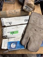 Boxes of welding rods from Akeloys and Achlands Limited, different sizes visible including 5mm x 300mm and 2.5mm x 300mm, arranged on a wooden surface with a protective leather glove.